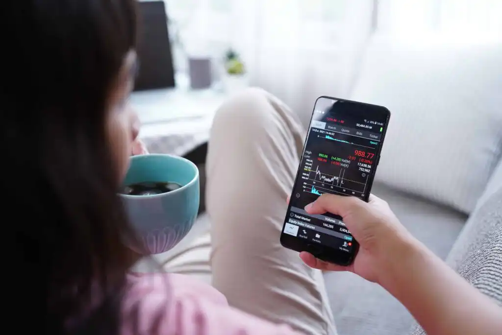 A person sits on a couch, holding a mug and looking at a smartphone displaying a stock trading app with charts and stock data. A laptop is on a table in the blurry background.