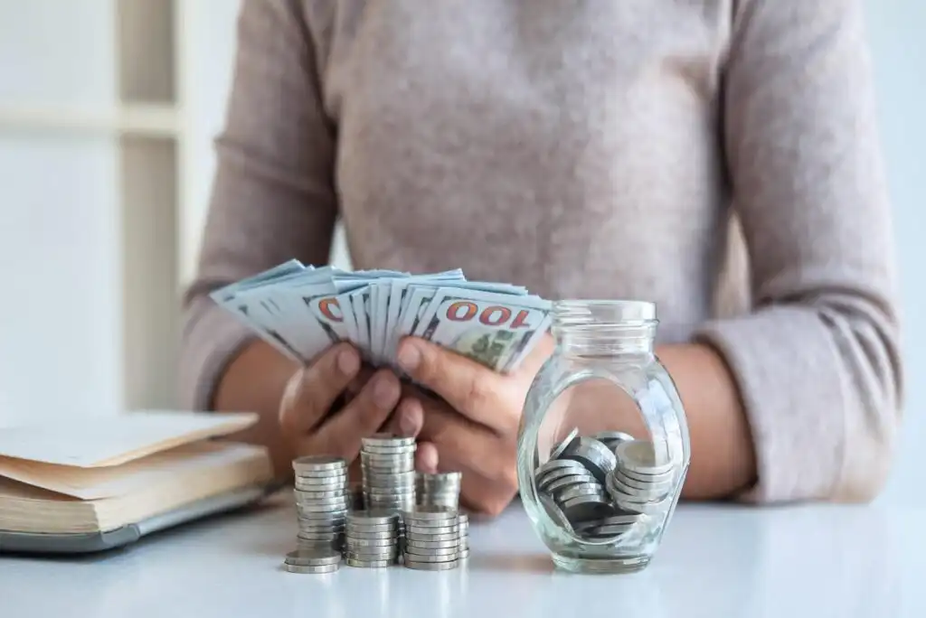 A person counts a stack of $100 bills at a table with several piles of coins and a small glass jar filled with coins in the foreground.