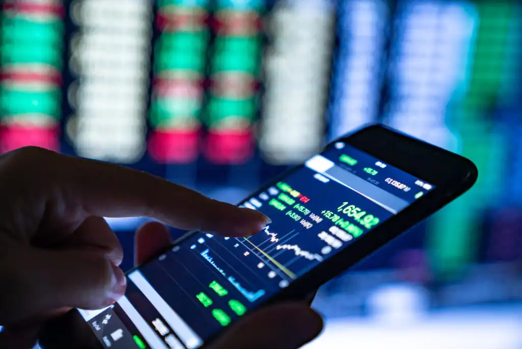 A person uses a smartphone displaying a stock market app with charts and numbers, while blurred stock prices in red and green are visible on a large screen in the background.