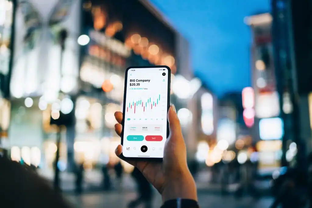 A person holds a smartphone displaying a stock trading app with a candlestick chart for “BIG Company.” The background shows a brightly lit city street at dusk, out of focus.