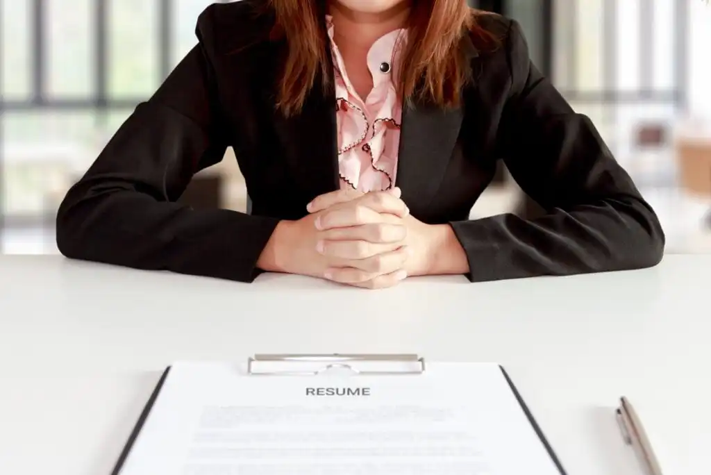 A woman in a business suit sits at a desk with her hands clasped, facing a resume on a clipboard that is in focus in the foreground. Her expression is not visible.