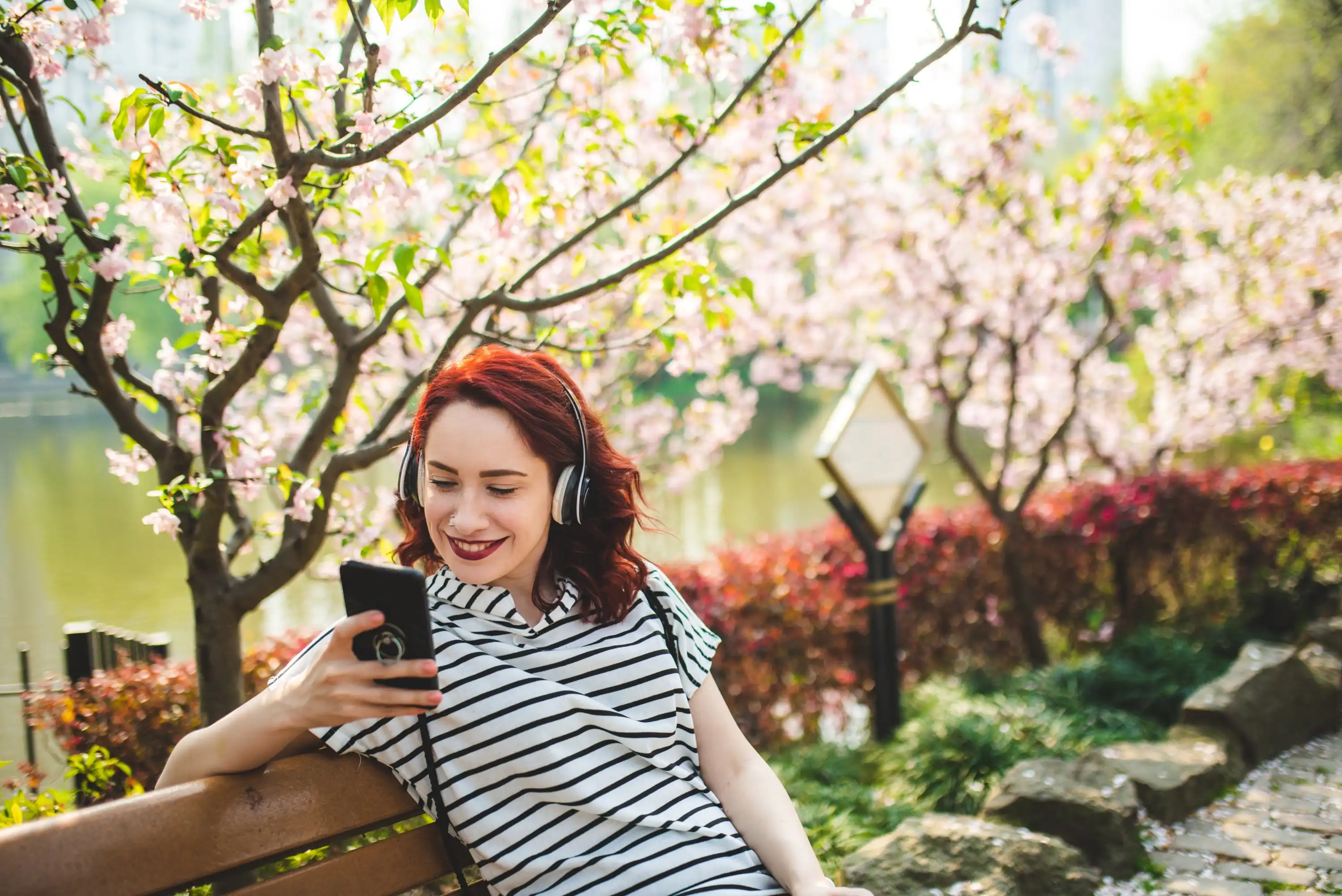 A young woman with red hair sits on a park bench, smiling while listening to headphones and looking at her phone. Pink-blossomed trees and a pond are in the background, creating a peaceful spring scene.