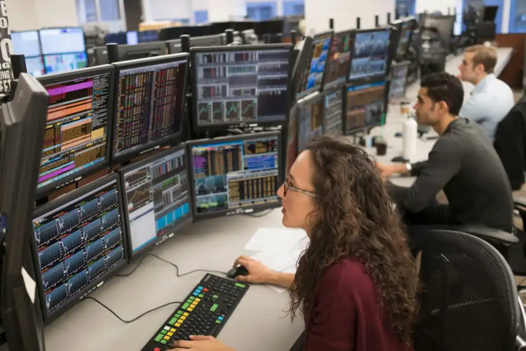 Three people sit at desks in an office, each working at computer stations with multiple large monitors displaying financial charts, graphs, and data. The focus is on a woman with curly hair in the foreground.