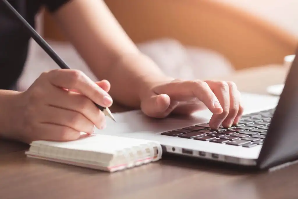 A person writes in a notebook with one hand while using the other hand to type on a laptop keyboard, working at a wooden desk in soft natural light.