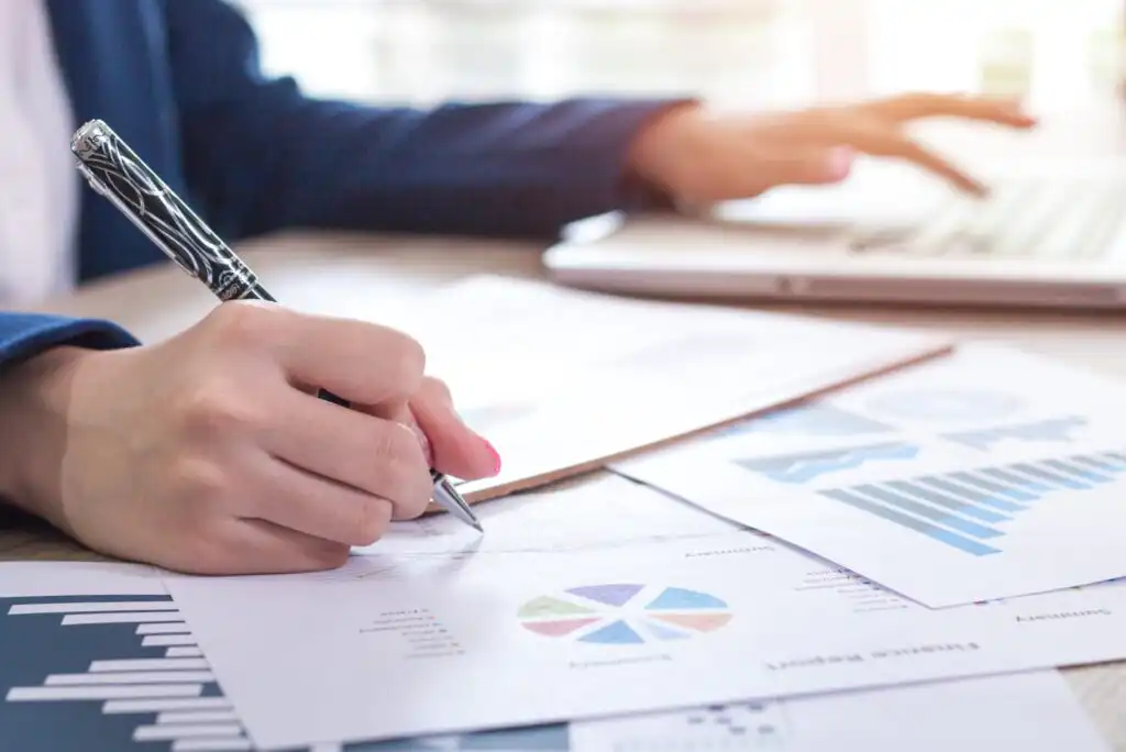 A person in business attire writes on financial documents with charts and graphs, while using a laptop on a desk in a bright office setting.