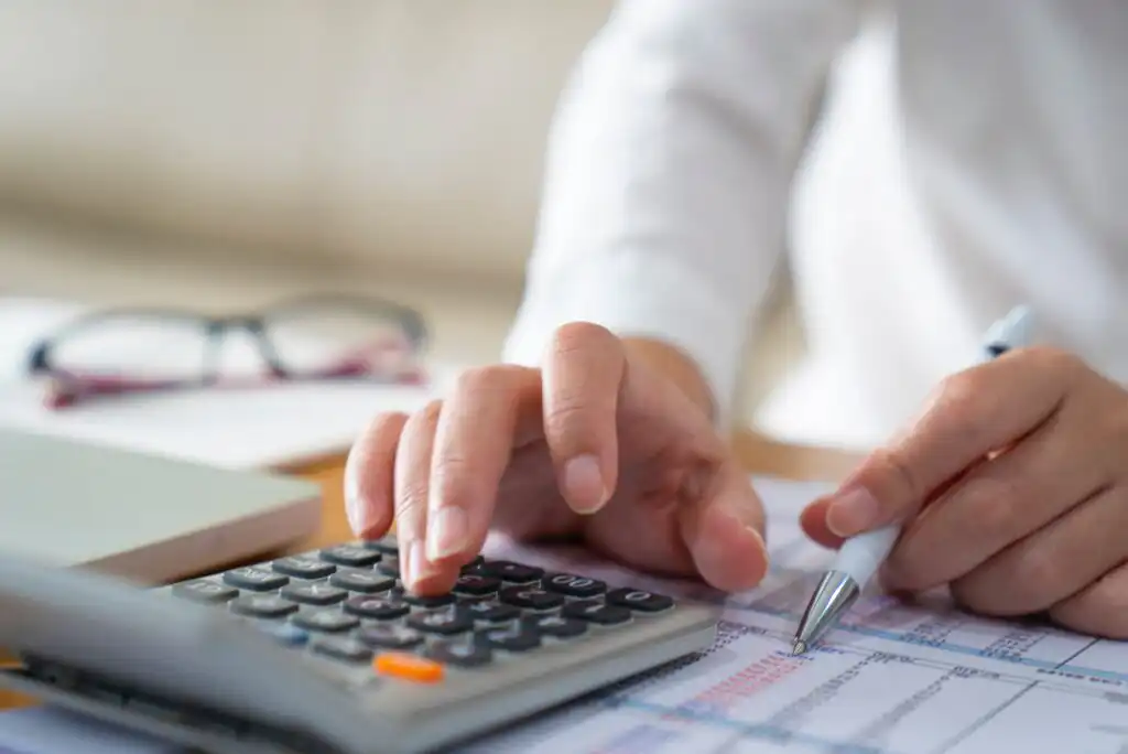 A person uses a calculator and holds a pen, working on financial documents at a desk. Glasses and papers are also visible in the background.