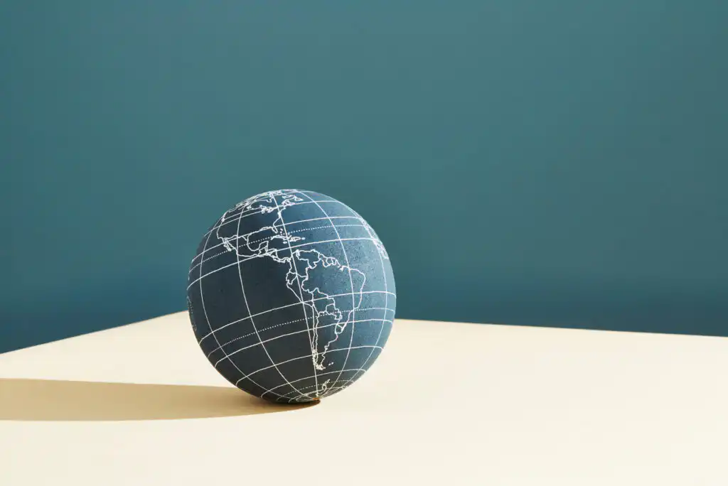 A globe with white grid lines and outlines of North and South America sits on a beige surface against a blue background, casting a shadow to the left.