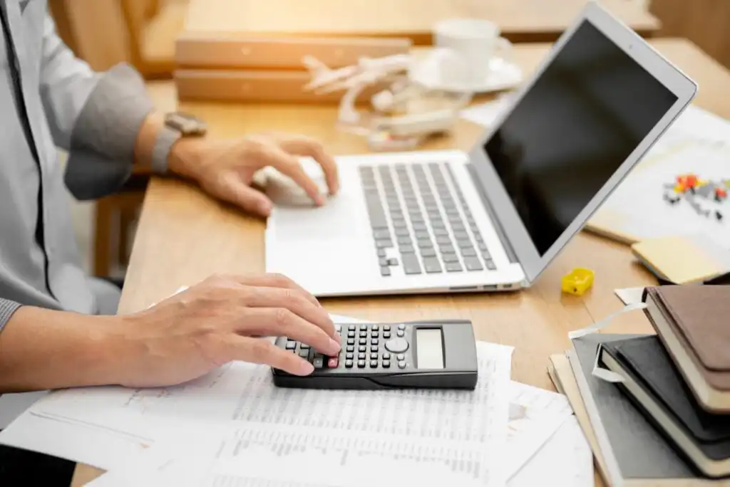 A person using a calculator and a laptop at a cluttered desk with papers, documents, and office supplies.