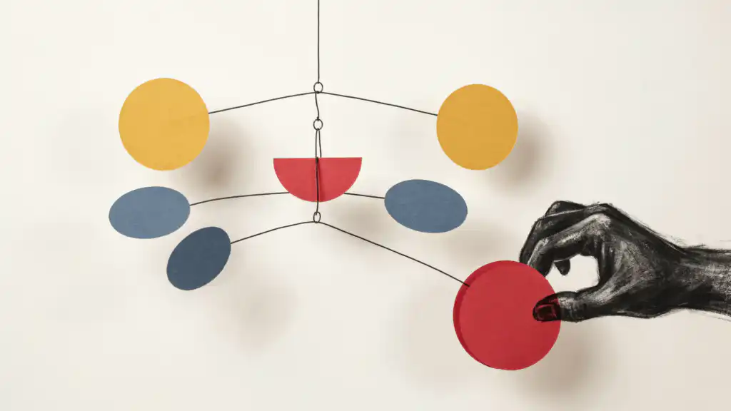 A charcoal hand grips a red disc on a balanced paper mobile, tipping its lowest arm while the arms above stay level