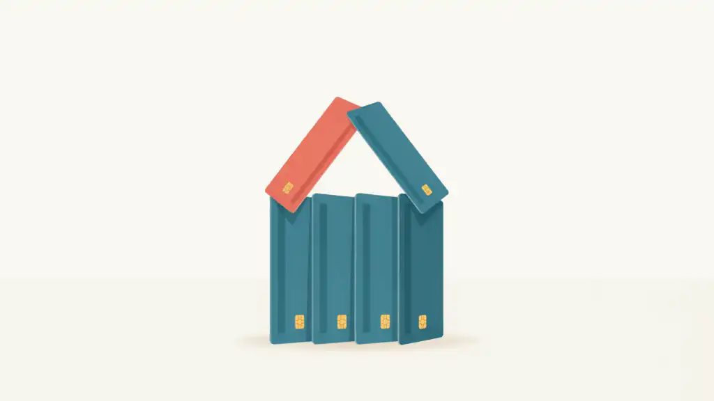 A small house built from teal credit cards with a coral card on its roof, balanced on a cream tabletop.
